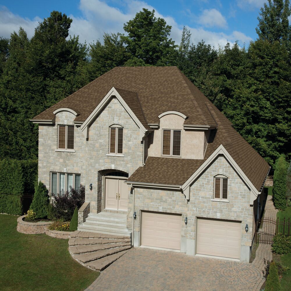 A two-story stone house with an Earthtone Cedar roof, two garage doors, and a paved driveway, nestled in the picturesque surroundings of Cambridge against a backdrop of trees and blue sky.