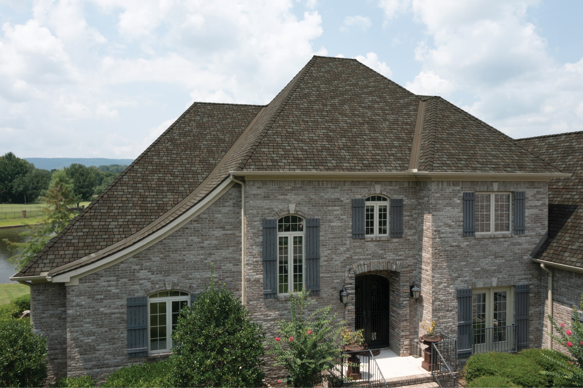A two-story brick house with a steep, multi-gabled roof finished in Harvest Slate, large windows with shutters, and a central entrance. A serene lawn and trees enhance its Royal Estate allure in the background.