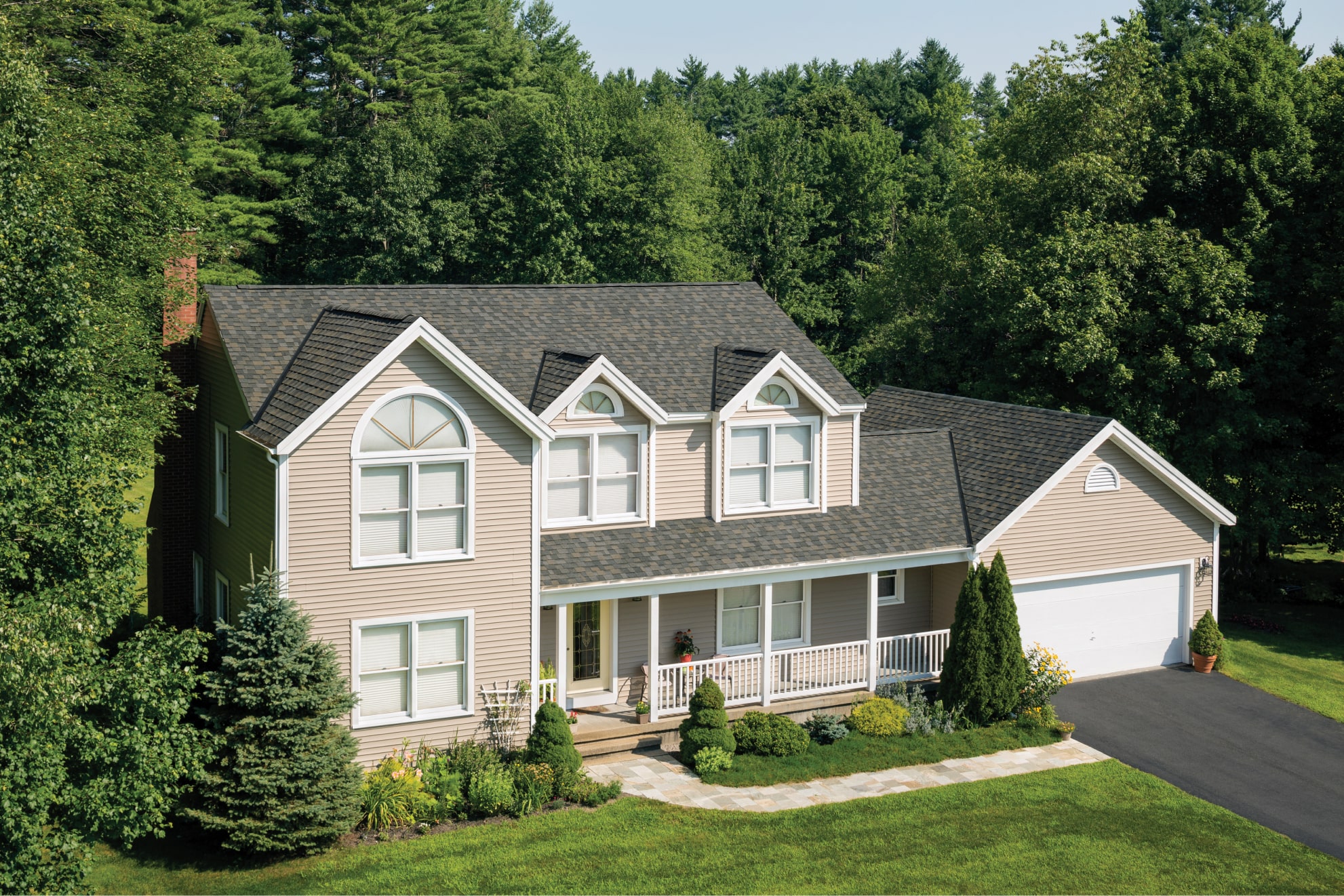 The two-story beige house, reminiscent of a Biscayne retreat, features a dark roof and is surrounded by trees and a manicured lawn. A white garage, paved driveway, and small front porch complete this scene of suburban tranquility.