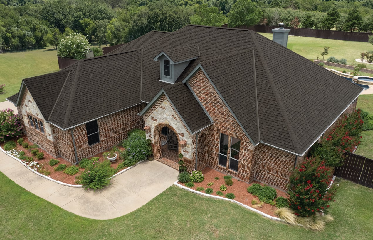 Aerial view of a large brick house with a dark roof, nestled near Cambridge. The property boasts a well-maintained lawn and landscaping, complemented by the natural beauty of driftwood accents around a nearby pool and wooded area.