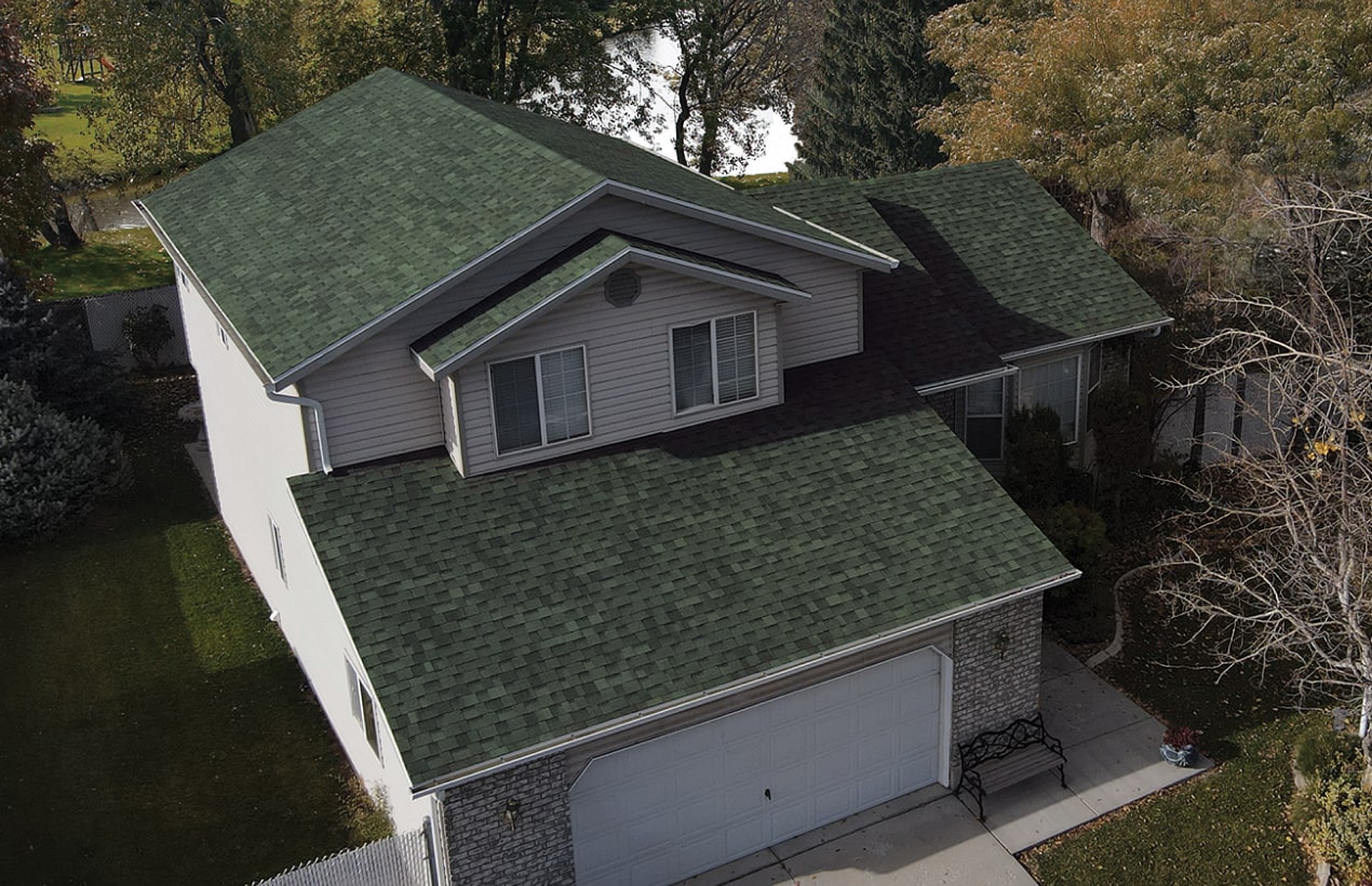 An aerial view reveals a charming two-story house with an emerald green shingle roof, white siding, and brick accents, nestled gracefully amidst a dynasty of towering trees.
